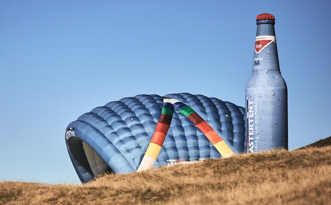 Two inflatable advertising media are located side by side on a meadow: model Kokon and model Bottle. Both in the CI colours of MASTERTENT and with a logo print.