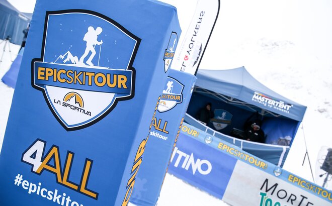 In primo piano si vedono dei pali con il logo dell evento Epic Ski Tour e delle transenne con della pubblicità. Nello sfondo c' è un gazebo pieghevole MASTERTENT blu. 
