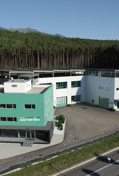 Das Firmengebäude Mastertent ist grün-grau. Im Hintergrund befindet sich ein Wald. 