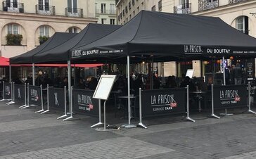 On the outdoor area of a restaurant there are standing three big, black folding gazebos with the La Prison label. Under the tents there are many tables and chairs.