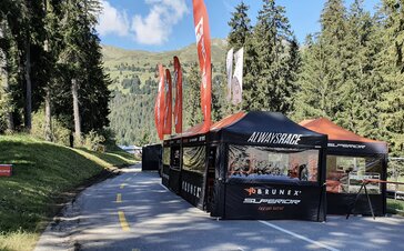 On a road there are several black folding gazebos with orange flags and roofs of a racing team. The road is surrounded by woods.