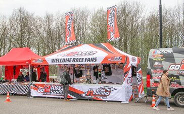 Ein weißer Faltpavillon mit Dachflaggen steht bei einem Motorsportevent. Darunter werden Werbeartikel verkauft.