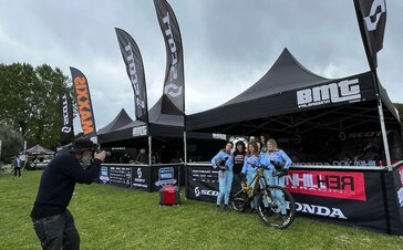 Three 5x5 m folding gazebos stand on a lawn. They are black and personalised with white logos from Scott, BMT and Honda. All three folding gazebos have half-height sidewalls and printed rear walls, as well as large, personalised flags on the roof. The racers and a photographer are in the foreground.
