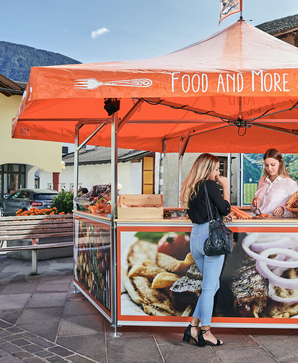 A semi-permanent orange and printed pavilion event tent structure set up to sell produce at a market.