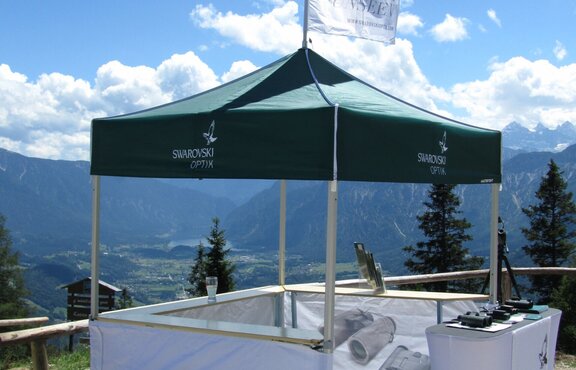 A green 5x5 canopy tent with custom printing, sidewalls, counters and a peak flag serves as an outdoor marketing stand. 