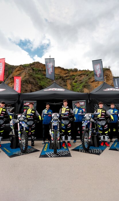 Auf dem Bild sind drei schwarze Faltpavillons mit Fahnen zu sehen. Die Zelte sind mit dem Logo des Motorradteams bedruckt. Das Team steht vor den Zelten.