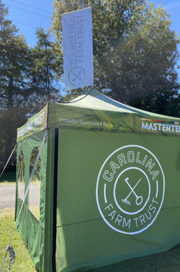 A green, 10x10ft Mastertent Canopy Tent printed with Carolina Farm Trust branding. Equipped with tent sidewalls and a peak flag.
