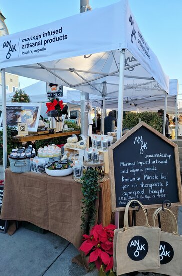 A white 10x5ft Mastertent canopy tent set up at a market to sell Anako moringa infused products at a farmer's market.