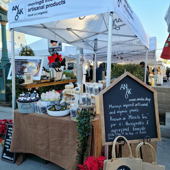 A white 10x5ft Mastertent canopy tent set up at a market to sell Anako moringa infused products at a farmer's market.