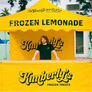 A man standing behind a counter for a yellow, 10x10ft lemonade tent with a scalloped roof, custom printing, and yellow sidewalls.