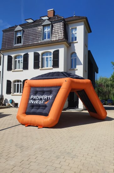 Branded black and orange tripod advertising inflatable in front of scenic house. 