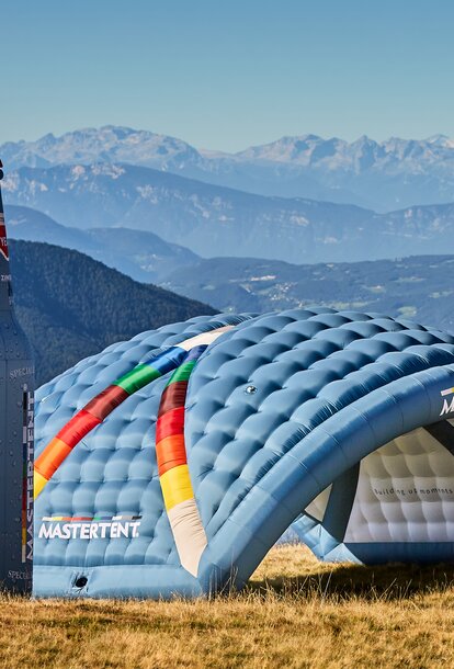 Three large inflatable structures in a field on a sunny day with mountains in the background. On the far left, a tall, blue and red bottle-shaped inflatable next to a curved, blue arch inflatable. On the right, a three-legged blue and white angular dome inflatable. 