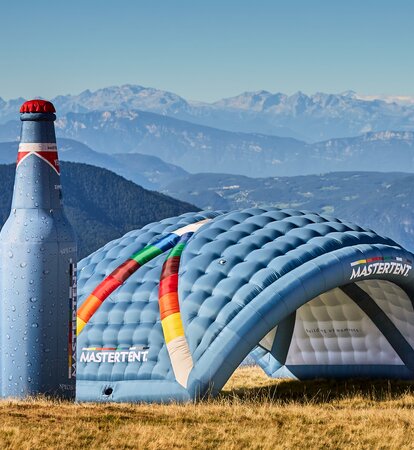 Three large inflatable structures in a field on a sunny day with mountains in the background. On the far left, a tall, blue and red bottle-shaped inflatable next to a curved, blue arch inflatable. On the right, a three-legged blue and white angular dome inflatable. 