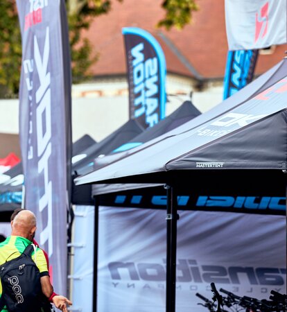 Closeup of black Mastertent canopy tents with awnings and flags at a cycling event with people in the background.