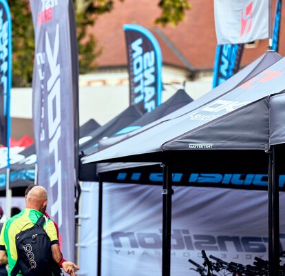 Closeup of black Mastertent canopy tents with awnings and flags at a cycling event with people in the background.