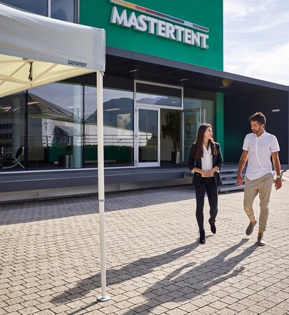 Man and woman talking and walking from modern, green building toward a white canopy tent on a sunny day