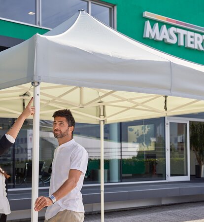 Man and woman talking and looking at a white canopy tent on a sunny day with a modern, green building in background