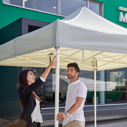Man and woman talking and looking at a white canopy tent on a sunny day with a modern, green building in background
