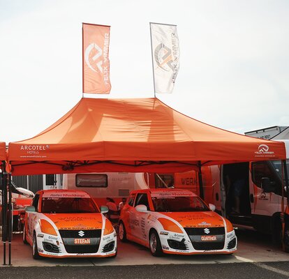 Large orange Mastertent tent with flags over two orange and white Suzuki racing cars 