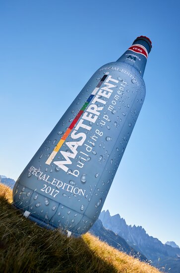 A large, blue inflatable in the shape of a classic glass bottle. Photographed at an angle with a mountain landscape in the background.