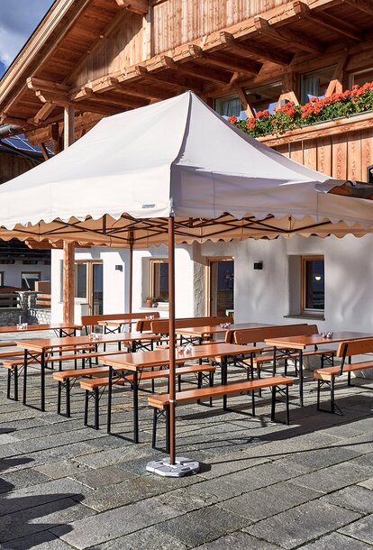 A large Mastertent canopy tent with white roof and brown frame set up outside of a wooden mountain lodge with rows of tables and benches underneath.