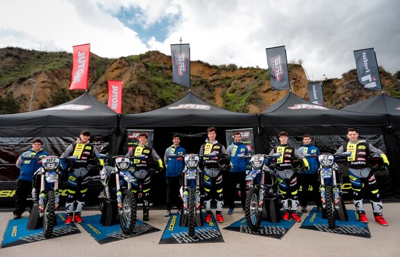 The picture shows three black folding gazebos with flags. The gazebos are printed with the logo of the motorbike team. The team is standing in front of the tents.