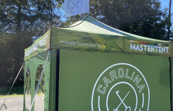 A green, 10x10ft Mastertent Canopy Tent printed with Carolina Farm Trust branding. Equipped with tent sidewalls and a peak flag.