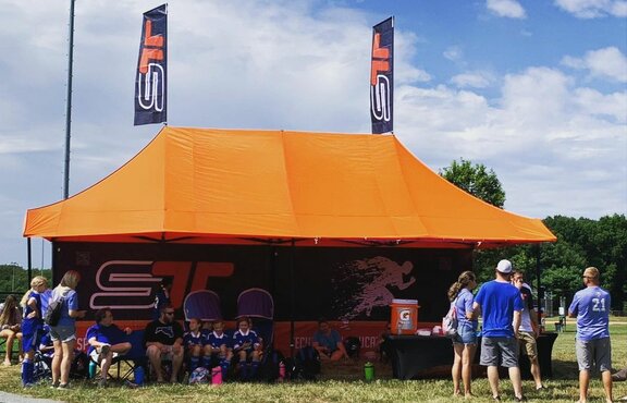 An orange Mastertent canopy tent with awnings, flags, and printed back wall set up at a sporting event for team concessions and shelter.