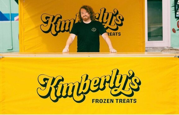 A man standing behind a counter for a yellow, 10x10ft lemonade tent with a scalloped roof, custom printing, and yellow sidewalls.