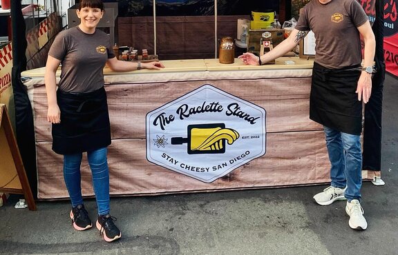 Two people stand in front of a Mastertent 10x10 custom printed canopy tent selling food at a market. 