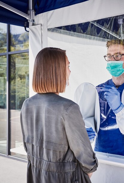 The employee inside the tent performs a smear test on the woman in front of it. He wears a mouth guard and gloves.