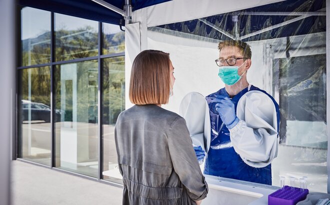 The employee inside the tent performs a smear test on the woman in front of it. He wears a mouth guard and gloves.