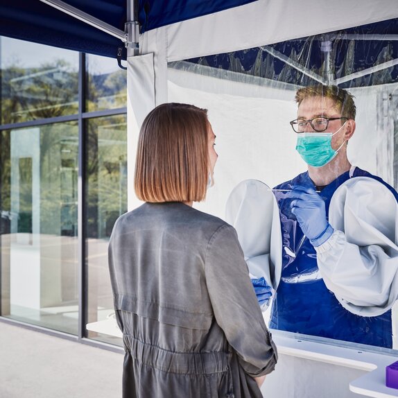 The employee inside the tent performs a smear test on the woman in front of it. He wears a mouth guard and gloves.