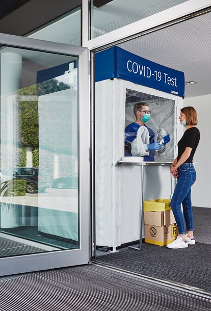 Corona test cabin at the entrance. The woman is standing in front of it and getting herself tested.