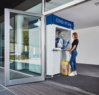 Corona test cabin at the entrance. The woman is standing in front of it and getting herself tested.