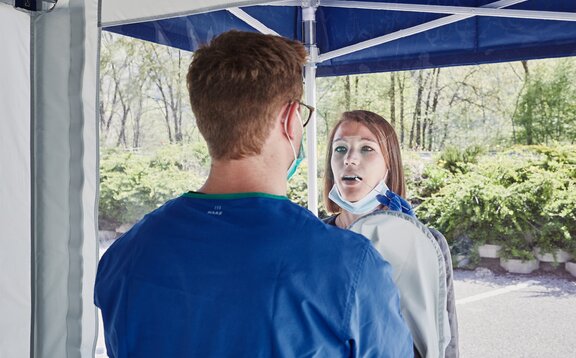 Der Mitarbeiter im Zeltinneren führt einen Abstrich durch die im Zelt integrierten Handschuhe durch. Die Frau befindet sich vor der Testkabine.