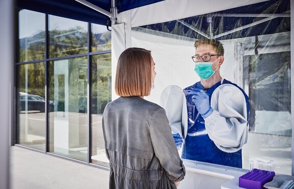 Der Mitarbeiter im Zeltinneren führt einen Abstrich bei der Frau davor durch. Er trägt einen Mundschutz und Handschuhe.