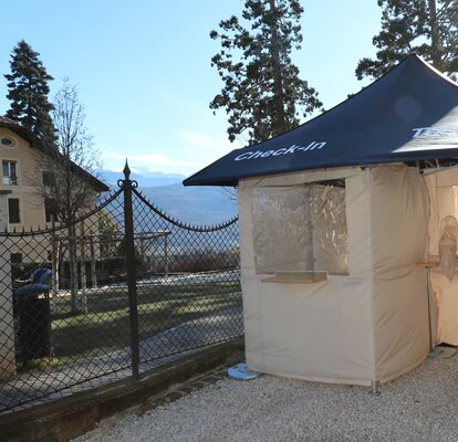 Il centro test Covid-19 si trova di fronte alla farmacia Kaltern nella Goldgasse. Il tetto è blu, le pareti laterali grigie e il sole splende nel cielo.