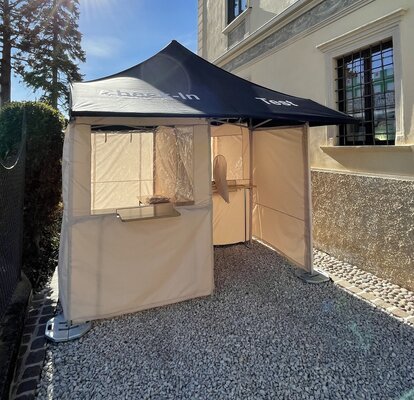 Die Covid-19 Serien-Test-Station steht vor der Apotheke Kaltern in der Goldgasse. Das Dach ist blau, die Seitenwände grau und am Himmel strahlt die Sonne.