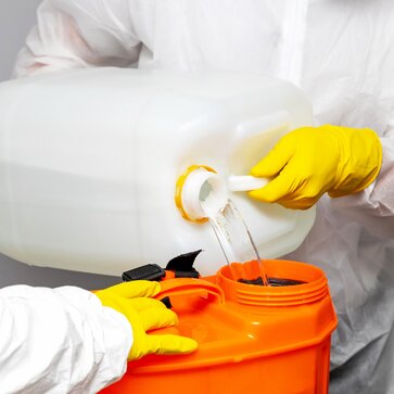Il disinfettante viene travasato da un contenitore bianco a uno arancione. La bomboletta è tenuta da due persone che indossano indumenti protettivi bianchi e guanti protettivi gialli. 