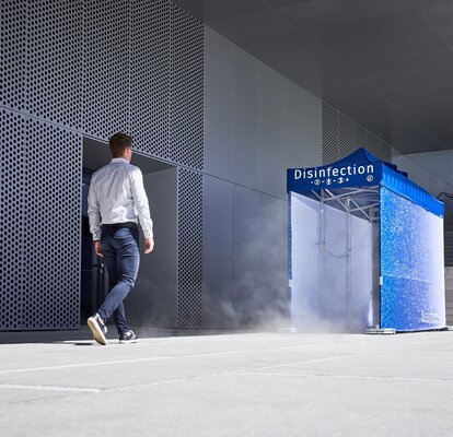 Un uomo sta per entrare nel tunnel di disinfezione blu, che è già pronto a spruzzare l'uomo dalla testa ai piedi e quindi a disinfettarlo. Il tunnel di disinfezione si trova accanto a un edificio grigio.