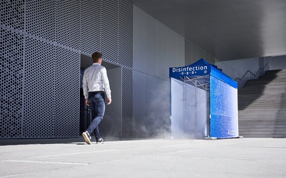 Un uomo sta per entrare nel tunnel di disinfezione blu, che è già pronto a spruzzare l'uomo dalla testa ai piedi e quindi a disinfettarlo. Il tunnel di disinfezione si trova accanto a un edificio grigio.