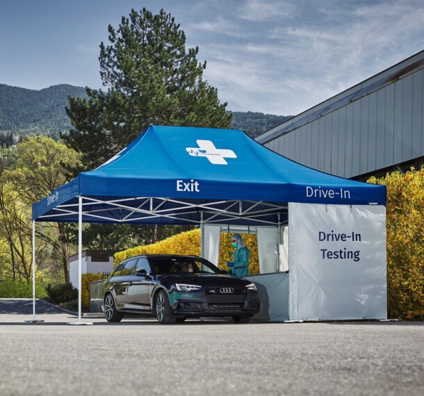 The black car drives to the drive-in test station. The drive-in test station is in a parking lot.