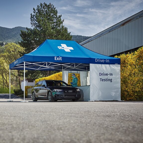 Das schwarze Auto fährt zur Drive-In Teststation. Die Drive-In Teststation steht auf einem Parkplatz.