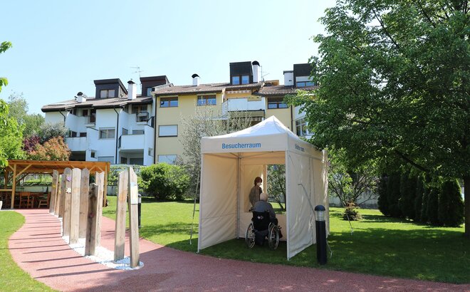 A mobile visitor room for the old people's home. The elderly gentleman sits on one side of the room and behind him stands the woman. Both are separated by a transparent wall.