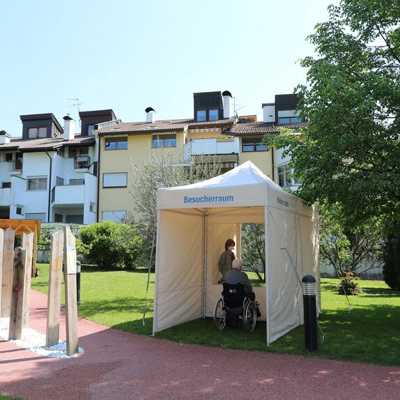 Ein mobiler Besucherraum für das Altersheim. Der ältere Herr sitzt auf einer Seite des Raums und dahinter steht die Frau. Beide sind durch eine transparente Wand getrennt.