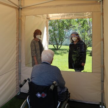 Besucherraum von innen. Der Senior im Rollstuhl sitzt vor der transparenten Wand. Dahinter stehen 2 Frauen mit Mundschutz.