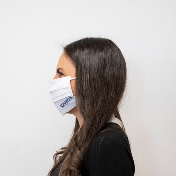 Eine Frau trägt eine waschbare Stoffmaske. Auf der Seite der weißen Maske ist ein blaues Firmenlogo abgebildet. 