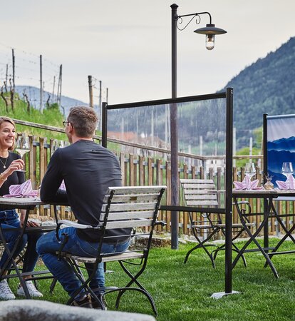 Trennwand steht zwischen den Tischen im Gastgarten. Das Paar sitzt an einem gedeckten Tisch. Dahinter sieht man noch eine bedruckte Trennwand.