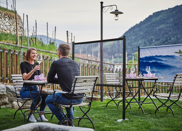 Trennwand steht zwischen den Tischen im Gastgarten. Das Paar sitzt an einem gedeckten Tisch. Dahinter sieht man noch eine bedruckte Trennwand.